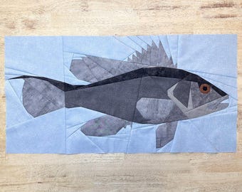 May include: A geometric fabric art piece featuring a stylized fish in shades of gray and black, set against a light blue background. The fish has a detailed fin and a brown eye. The artwork is likely a quilt block or a similar textile creation.