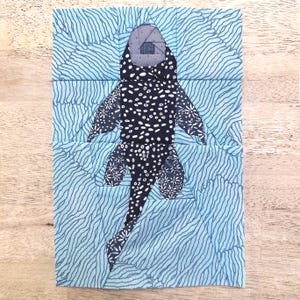 Puede incluir: Una pieza de arte textil que presenta una criatura estilizada de lunares blancos y negros con detalles florales, sobre un fondo azul claro ondulado. La obra de arte es rectangular y parece ser un bloque de colcha.