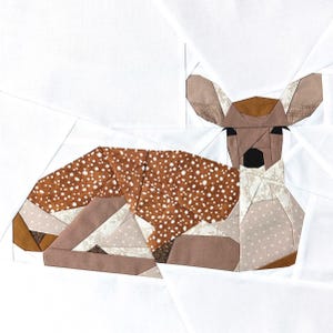 Op de afbeelding: Een patchwork quiltblok met een liggend, bruin-wit gevlekt hert. Het hert is gemaakt van verschillende stofstukken die in een geometrisch patroon aan elkaar zijn genaaid.