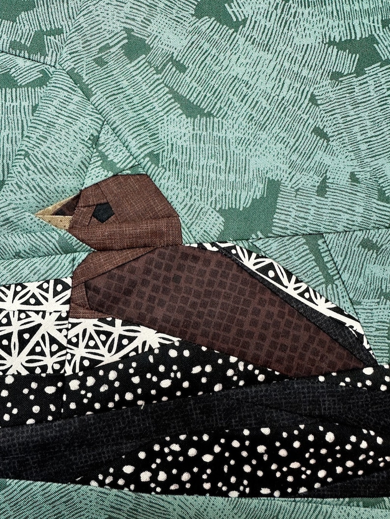 Peut inclure: Gros plan d'une courtepointe d'oiseau en tissu. L'oiseau est brun avec un motif g&eacute;om&eacute;trique, sur un fond vert avec un motif textur&eacute;. L'oiseau est perch&eacute; sur un nid en tissu &agrave; motifs noir et blanc.