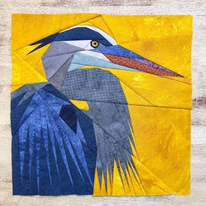 May include: A blue and yellow patchwork quilt featuring a stylized heron with a long beak. The heron's head is turned to the right and its eye is visible. The quilt is made of fabric squares sewn together to create a geometric pattern.