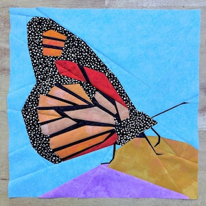 May include: A colorful paper pieced butterfly with black, orange, red, and white fabric. The butterfly is perched on a purple and gold fabric flower.