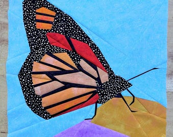 May include: A colorful paper pieced butterfly with black, orange, red, and white fabric. The butterfly is perched on a purple and gold fabric flower.