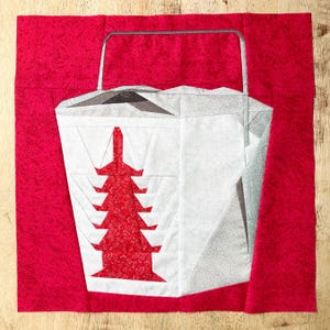 May include: A quilted block featuring a white takeout container design with a red tree pattern. The container has a grey handle and is set against a red background. The design is made of fabric pieces.