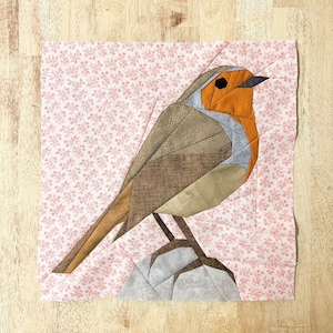 Puede incluir: Un bloque de colcha de patchwork con un petirrojo de color marrón, naranja y gris. El pájaro está posado en una rama con un fondo floral rosa.