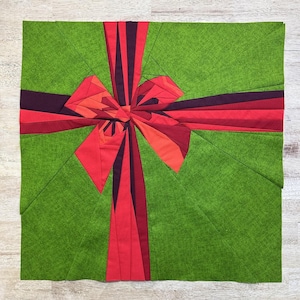 May include: A green fabric square with a red and burgundy bow in the center. The bow is made of multiple fabric pieces sewn together to create a 3D effect.