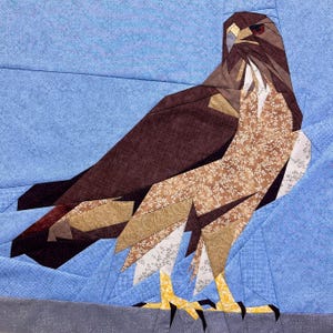 Pode incluir: Um bloco de colcha de retalhos marrom e branco representando um falcão com um olho vermelho. O falcão está em pé sobre uma superfície cinza em um fundo azul.