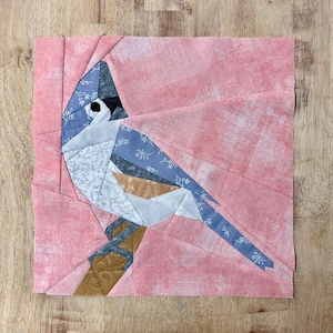May include: A paper pieced quilt block featuring a bluebird perched on a branch. The bird is made of various shades of blue, white, and brown fabric. The background is a soft pink with a subtle white pattern.