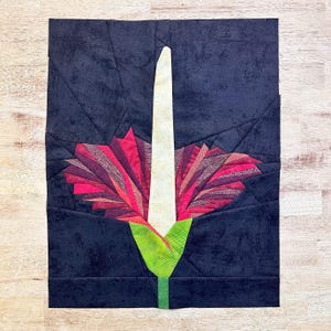 May include: A fabric art piece featuring a stylized flower. The flower has a white central column, surrounded by layered petals in shades of red and pink. The flower sits on a green stem against a dark blue background.