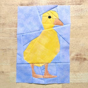 Op de afbeelding: Een stoffen blok met een geometrisch ontwerp van een gele eend. De eend heeft een zwart oog, een oranje snavel en poten, tegen een lichtblauwe achtergrond met een stippenpatroon. Voor quilten of knutselen.