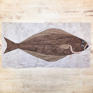 May include: A fabric art piece featuring a brown fish with a flat body and a pointed tail. The fish is constructed from various shades of brown fabric, with a textured appearance. The background is a light gray color, and the artwork is displayed on a wooden surface.