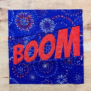 May include: A red fabric applique of the word "BOOM" on a blue background with a white and red firework pattern.