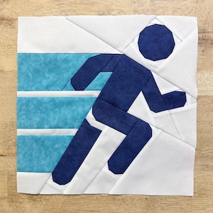 May include: A white fabric quilt block with a blue running figure design. The figure is made of multiple fabric pieces sewn together. The background is white with a blue and white striped pattern.