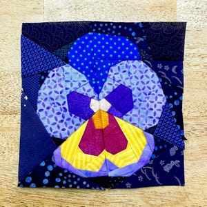 Puede incluir: Una obra de arte textil que representa un pensamiento estilizado. La flor está compuesta por formas geométricas azules, moradas, amarillas y blancas. El fondo es un patchwork de varias telas azules estampadas. La obra de arte es cuadrada y descansa sobre una superficie de madera clara.