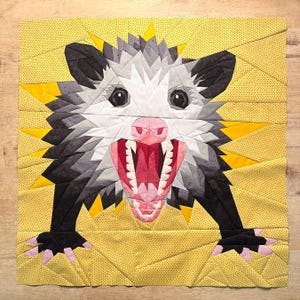 May include: A fabric artwork depicting an opossum in a geometric style. The opossum has a white face, grey and black fur, and a pink mouth. The background is yellow with a dotted pattern, and yellow starburst shapes surround the head.