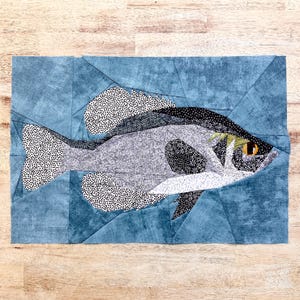 May include: A patchwork quilt depicting a black and white fish with a yellow eye, swimming in a blue and green water background.