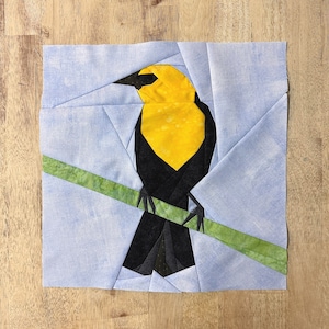 May include: A paper pieced quilt block featuring a yellow and black bird perched on a green branch. The bird has a yellow breast and black wings and tail. The background is a light blue fabric.