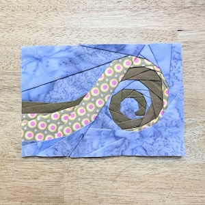May include: A blue, brown, and pink fabric quilt block with a spiral design. The spiral is made of fabric strips with a polka dot pattern.