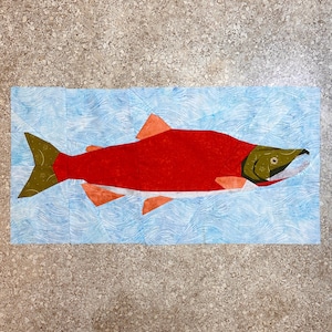 Puede incluir: Un salmón de tela roja y verde nadando en un océano de tela azul y blanco. El salmón está hecho de piezas de tela cosidas juntas para crear un efecto 3D.
