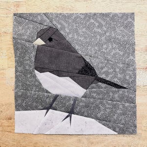 May include: A paper pieced quilt block featuring a black and white bird perched on a white snowdrift against a gray background with a floral pattern.