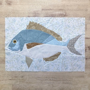 Puede incluir: Una obra de arte textil que representa un pez estilizado. El pez está compuesto por piezas de tela en tonos azules, marrones y blancos, con una cola estampada. El fondo es una tela azul claro con un estampado floral.