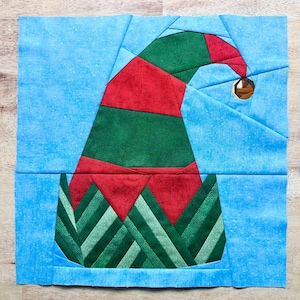 May include: A fabric block featuring a festive elf hat design. The hat is primarily red and green, with a gold-colored button detail. The background is a light blue, creating a holiday-themed quilt block.