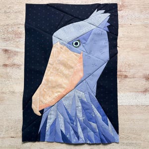 以下が含まれることがあります： ハシビロコウを描いた布製アート作品。鳥の頭と首は、青、グレー、ピーチ色の様々な生地で構成され、背景は黒です。鳥の目は小さな緑色の円です。