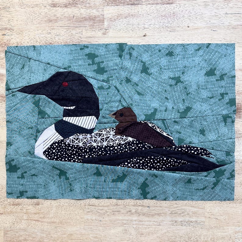 Peut inclure: Une &oelig;uvre d'art textile repr&eacute;sentant un huard et son petit. Le huard a une t&ecirc;te noire, une poitrine avec des motifs blancs et noirs, et un corps noir avec des pois blancs. Le petit est brun. Le fond est turquoise.