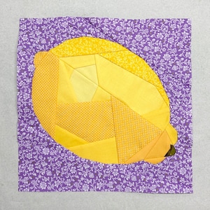 Puede incluir: Un bloque de colcha de retazos en forma de limón amarillo con un fondo floral morado.