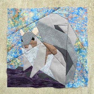 Può includere: Un'applique di tessuto grigio e bianco a forma di scoiattolo su uno sfondo di tessuto viola. Lo scoiattolo è composto da diversi pezzi di tessuto cuciti insieme per creare un effetto 3D. Il tessuto di fondo ha un motivo floreale.