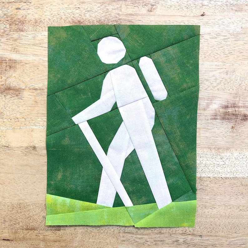 May include: A green and white fabric art piece depicting a person hiking with a backpack and walking stick. The figure is white against a green background, with a strip of green at the bottom representing grass. The artwork is made of fabric.