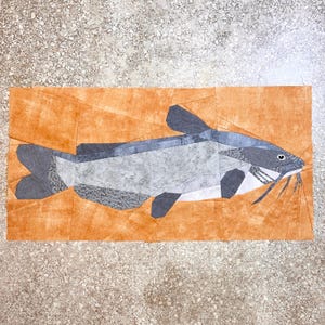 May include: A patchwork quilt depicting a catfish with a gray and white body and black fins. The catfish is on a brown background.