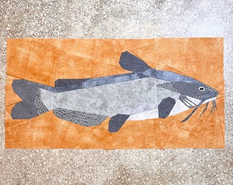 May include: A patchwork quilt depicting a catfish with a gray and white body and black fins. The catfish is on a brown background.