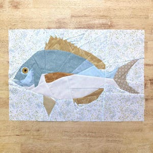 May include: A rectangular fabric quilt block featuring a pieced fish design. The fish is constructed from blue, white, and tan fabric segments with a yellow eye. The background is a light blue and white floral print fabric with visible seams.