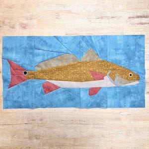 May include: A rectangular fabric quilt block featuring a pieced fish design. The fish has a golden-brown body, a white underbelly, and red fins with a circular spot on the tail. The background is a mottled blue fabric representing water.