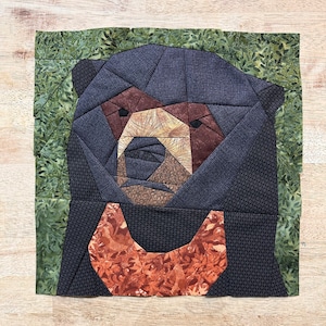 May include: A close-up of a black bear face made from fabric scraps. The bear's face is made up of different colored fabrics, including brown, black, and white. The bear's eyes are black and the nose is brown. The bear's mouth is open and the teeth are visible. The bear's face is surrounded by a green fabric background.