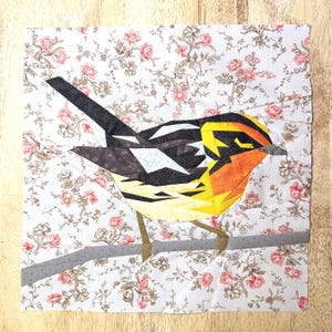 May include: A fabric quilt block featuring a pieced bird design with black, white, yellow, and orange geometric sections. The bird is perched on a gray branch against a white background patterned with small pink floral sprigs and brown vines.