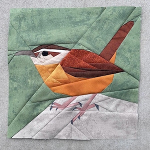 Könnte beinhalten: Ein Stoff-Quiltblock mit einem Zaunkönig-Design. Der Vogel hat braune, orange und weiße Federn, einen langen Schnabel und ein dunkles Auge. Der Hintergrund ist ein strukturierter grüner Stoff, und der Vogel sitzt auf einer grauen Oberfläche.