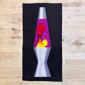 Puede incluir: Un bloque de colcha de lámpara de lava de colores con un flujo de lava morado, rojo, naranja y amarillo. La lámpara está rodeada de un fondo negro.