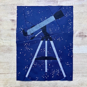 May include: A fabric art piece featuring a telescope against a dark blue starry night sky. The telescope is black and light blue, with white accents on the tripod legs. White stars are scattered across the background.