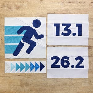 May include: A white fabric quilt block with a blue running figure, a blue fabric block with the number "13.1", a blue fabric block with the number "26.2", and a blue and white fabric block with a row of blue arrows.
