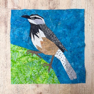 May include: A patchwork quilt block featuring a bird perched on a branch. The bird is made of fabric scraps in shades of brown, white, black, and gray. The branch is made of green fabric with a leaf pattern. The background is a blue fabric with a floral pattern.