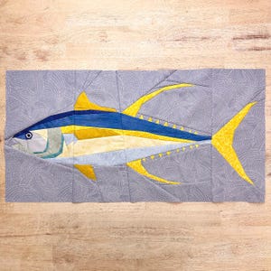 Puede incluir: Un bloque de edredón de tela rectangular con un diseño de atún. El pez está construido con segmentos de tela azul, amarilla y gris claro. El fondo consiste en una tela gris con un patrón circular blanco.