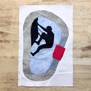 May include: A paper pattern for a quilt block featuring a climber in a rock climbing wall. The pattern is made of white paper with grey and blue fabric pieces. The climber is black and the wall is grey with a red accent.