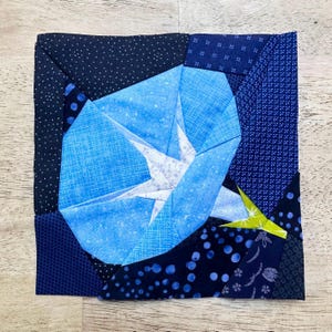 May include: A square fabric block featuring a blue flower design. The flower is a light blue with white accents, surrounded by various shades of blue fabric with different patterns. A small section of green is visible.