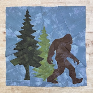 Peut inclure: Une silhouette de Bigfoot marron marchant dans une scène de forêt bleue et verte. Le Bigfoot est fait de tissu et est un bloc de courtepointe assemblé en papier.