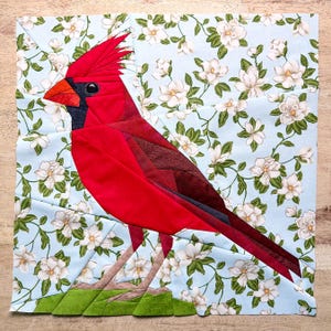 Op de afbeelding: Een quiltblok met een levendige rode kardinaalvogel. De vogel staat tegen een lichtblauwe achtergrond met witte bloempatronen. De kardinaal heeft een prominente kuif, zwarte accenten en zit op een groene basis. Het quiltblok is vierkant.