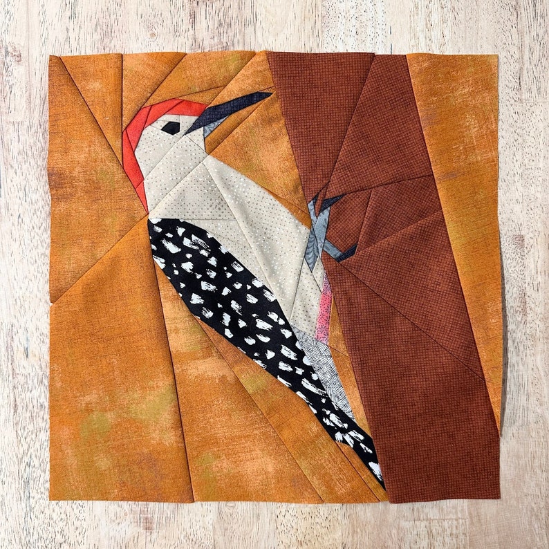 May include: A patchwork quilt block featuring a red-breasted woodpecker perched on a tree branch. The block is made of brown, orange, and white fabric.