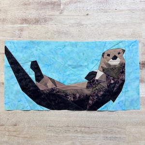Puede incluir: Una nutria marina de patchwork marrón y negro flota de espaldas en un fondo de agua azul y verde. La nutria está hecha de tela y es un bloque de colcha.