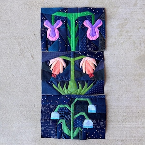 May include: A quilted floral design featuring three different flower types. The flowers are in shades of pink, purple, and blue, with green stems and leaves. The background is a dark blue fabric with white polka dots and other patterns.